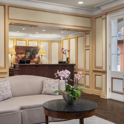 A hotel lobby with a reception desk, elegant seating, and decorative plants, featuring a view of a historical building outside the window.