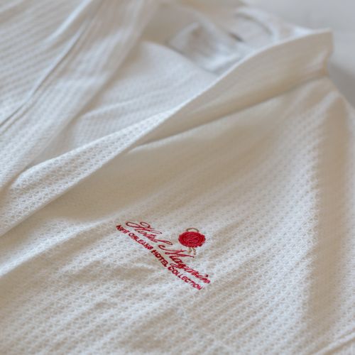 The image shows a white robe with red embroidery of a crown and text, likely a hotel or resort logo, on a bed.