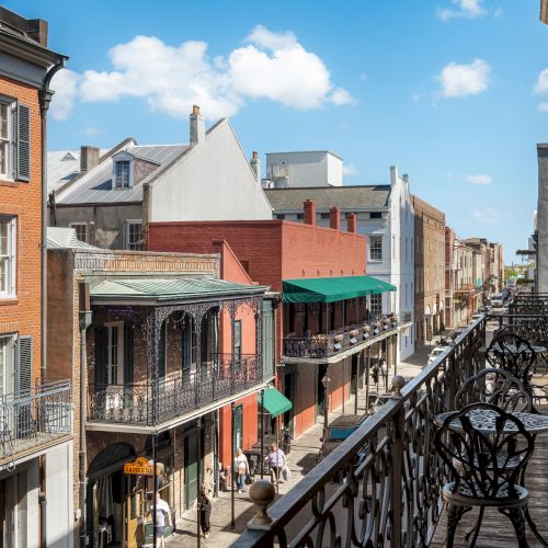 A street with historic buildings and wrought iron balconies on a sunny day, featuring a clear blue sky and some clouds.