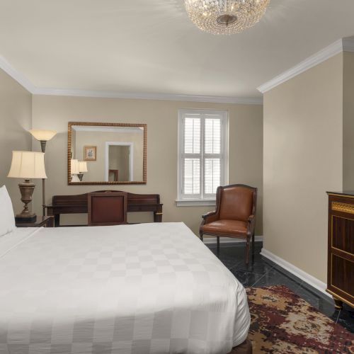 A hotel room with a bed, TV, chair, desk with mirror, lamp, and window, featuring beige walls and a rug underfoot, offers a cozy stay.