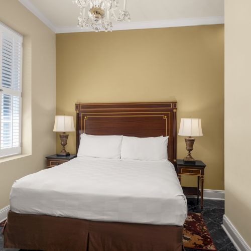 A cozy bedroom with a bed, two bedside lamps, and a framed picture on the wall. The room is warmly lit with beige walls and a window.