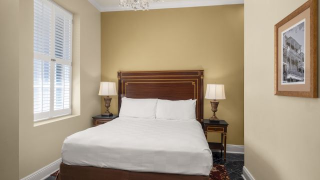 A neatly made bed with white linens and pillows, flanked by two nightstands with lamps, in a room with beige walls and a window with white blinds.