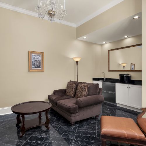A cozy living area with a brown sofa, armchair, coffee table, TV, and kitchenette, featuring elegant decor and a chandelier.