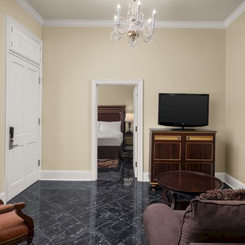 A living room with a chandelier, TV, sofa, armchair, and a glimpse of a bedroom through a doorway, showcasing elegance and comfort.