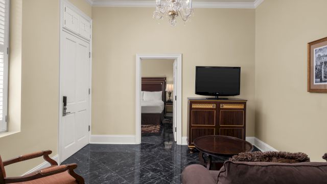 A wide-angle view of a hotel room's living area with a TV, sofa, armchair, and doorway to the bedroom.