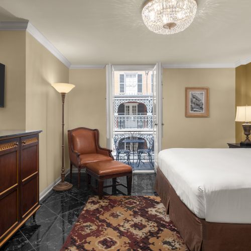 A hotel room with a bed, two lamps, a TV, armchair, dresser, and a window overlooking a balcony. A rug covers the polished floor.