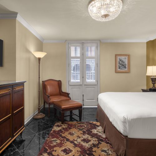 The image shows a hotel room with a bed, a chair, a dresser with a TV, a floor lamp, and a rug, all under a chandelier.