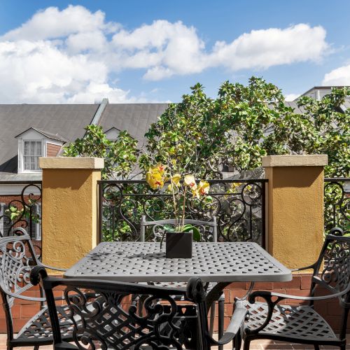 A small balcony with wrought iron furniture, a potted plant, and a view of trees and rooftops under a blue sky with clouds.