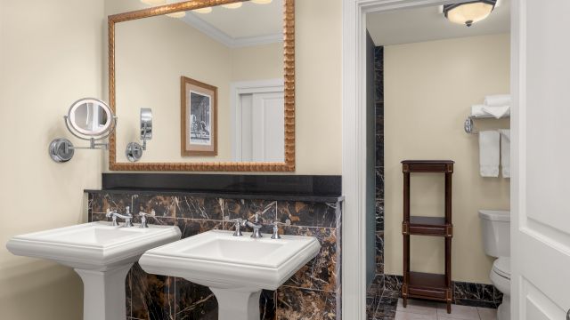 A bathroom with two white pedestal sinks, a large wall mirror, and a partially open door showing a toilet and shelving.