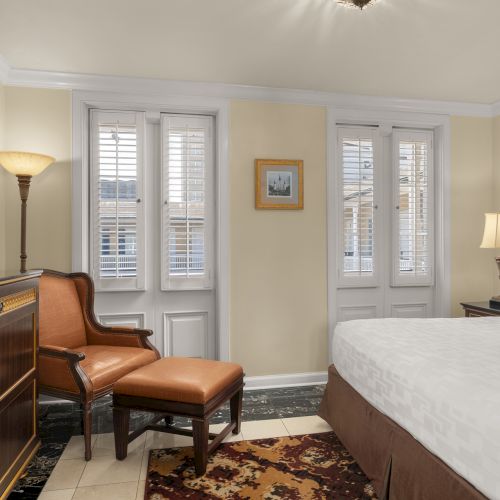 A cozy hotel room features a bed, TV, armchair with ottoman, lamps, and windows with shutters, creating a warm and inviting atmosphere.