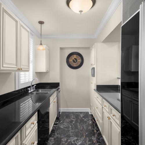 A modern kitchen with black countertops, white cabinets, and a wall clock. A sink, a fridge, and a pendant light are also visible.
