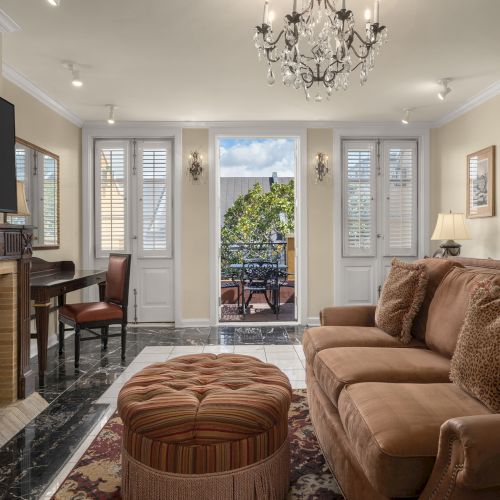 A cozy living room with a brown sofa, chandelier, TV, and fireplace, leading to a bright outdoor space through glass doors.