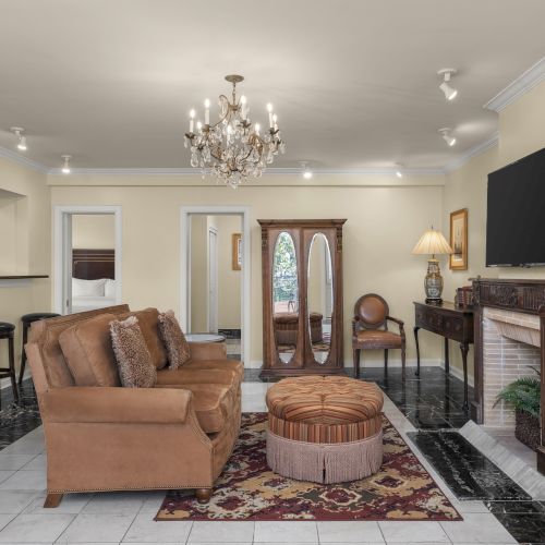 A cozy living room with a chandelier, sofa, oval ottoman, fireplace, TV, and elegant decor on a patterned rug, featuring warm lighting.