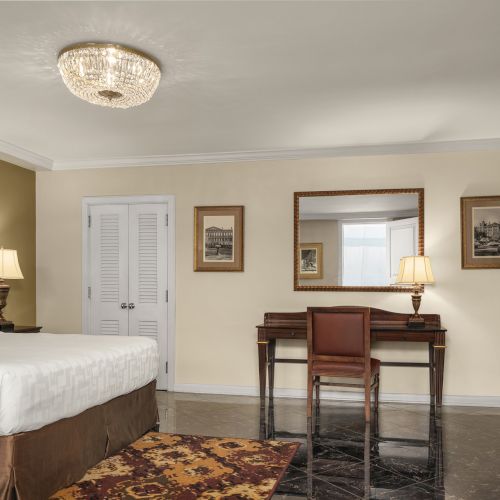 The image shows a hotel room with a bed, desk, chair, lamps, TV, mirror, and framed pictures on the walls, in a classic design.