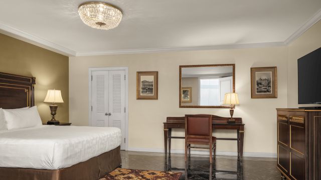 A wide view of a hotel room with a bed, desk, mirror, TV, and dresser.
