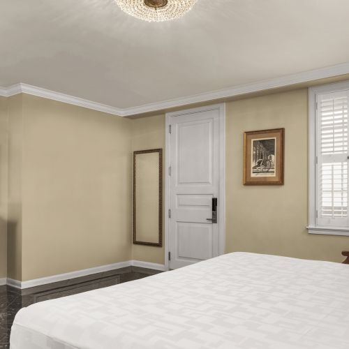 A bedroom with a bed, wooden TV stand, wall art, chair, window with shutters, and a closed door under a ceiling light.