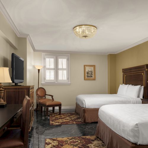 A hotel room with two beds, lamps, a TV, desk, and chairs. Decor is classic with neutral tones and a chandelier on the ceiling.