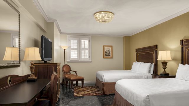 A hotel room with two beds, a desk, a TV, and a chandelier.