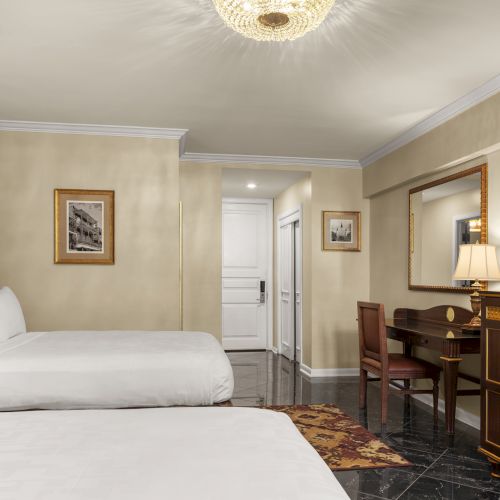 The image shows a hotel room with two beds, a desk with a lamp, a large TV, and elegant decor including a chandelier and framed artwork.