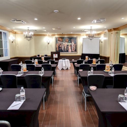 A conference room set up with tables, chairs, bottled water, and snacks, featuring chandeliers and a painting on the wall.