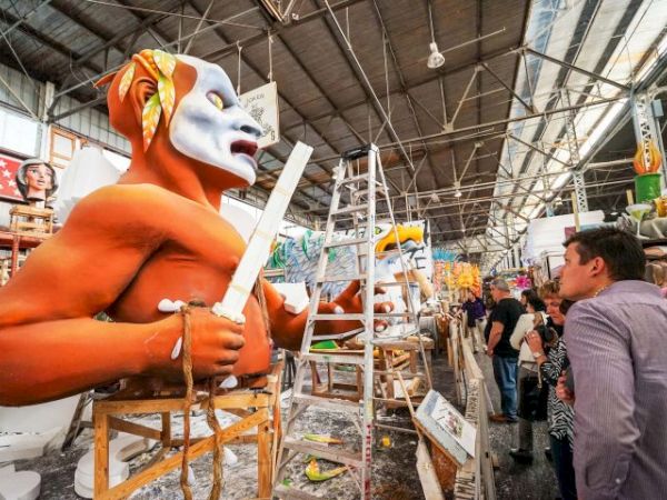 A craftsman builds large orange humanoid sculptures with white masks in a busy workshop, ladders and tools surround the scene, visitors watch.
