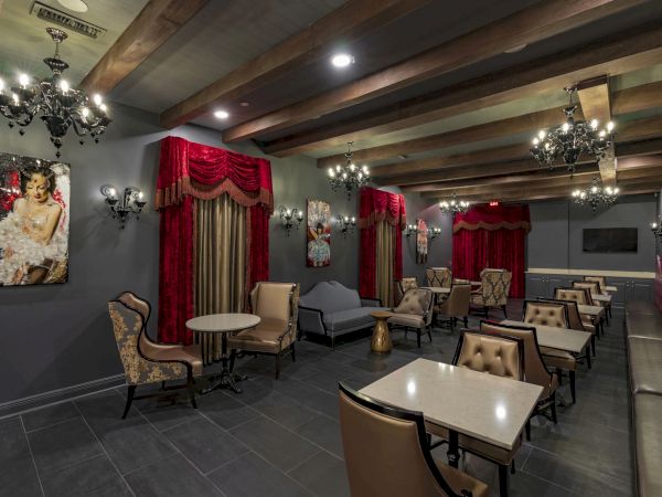 A stylish lounge or cafe with plush seating, red curtains, chandeliers, small tables, and mirrored walls for an intimate, upscale vibe.