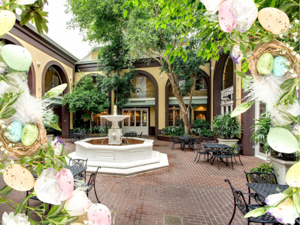 A sunlit courtyard with a central fountain, brick-paved ground, leafy trees, archways, and black metal caf&eacute; tables and chairs, framed by pastel egg decorations.