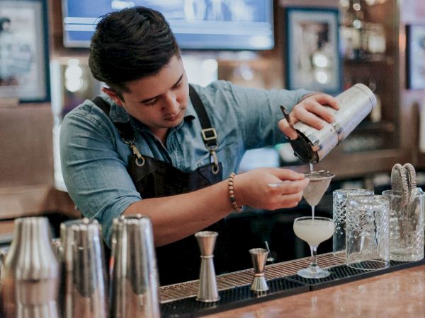 A bartender in a denim shirt and apron pours a cocktail through a strainer into a glass at a bar counter.