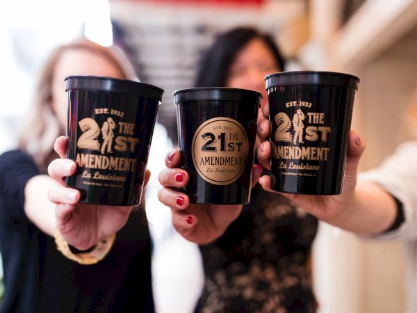 Three people holding black cups labeled "21st Amendment" from a place called La Louisiane, established in 1933.