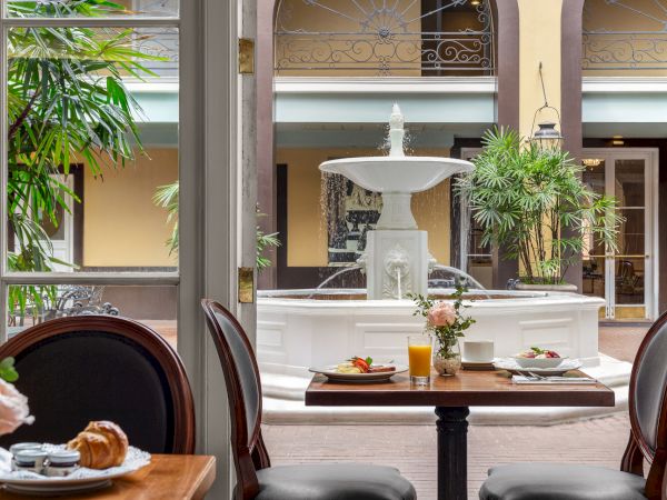 A dining area with breakfast on the table, elegant chairs, a fountain outside, and lush greenery in an open courtyard setting.
