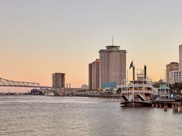 A waterfront city skyline with a wide river, high-rise buildings, and a docked riverboat in the golden-hour sky.