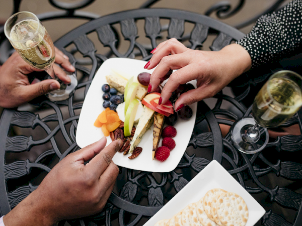 Hands and glasses clink over a platter of fresh veggies, fruit, and cheese on an outdoor patio table.