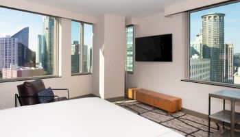 A modern hotel room with city views, featuring large windows, a bed, a TV, a chair, a table, and a wooden cabinet.