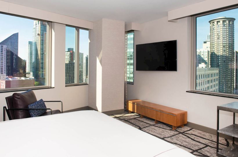 A modern hotel room with city views, featuring large windows, a bed, a TV, a chair, a table, and a wooden cabinet.