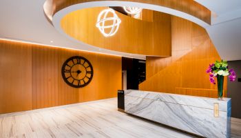 A modern lobby features a marble desk, a large wall clock, wooden accents, and a spiral light fixture.