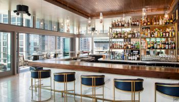 A modern bar with a sleek counter, blue and gold stools, and a well-stocked shelf, overlooking a cityscape through large windows.