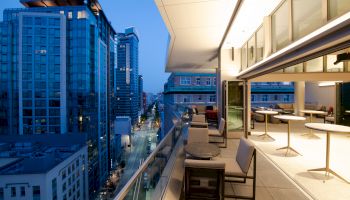 A modern city view from a balcony, with outdoor seating and buildings lit up at dusk.