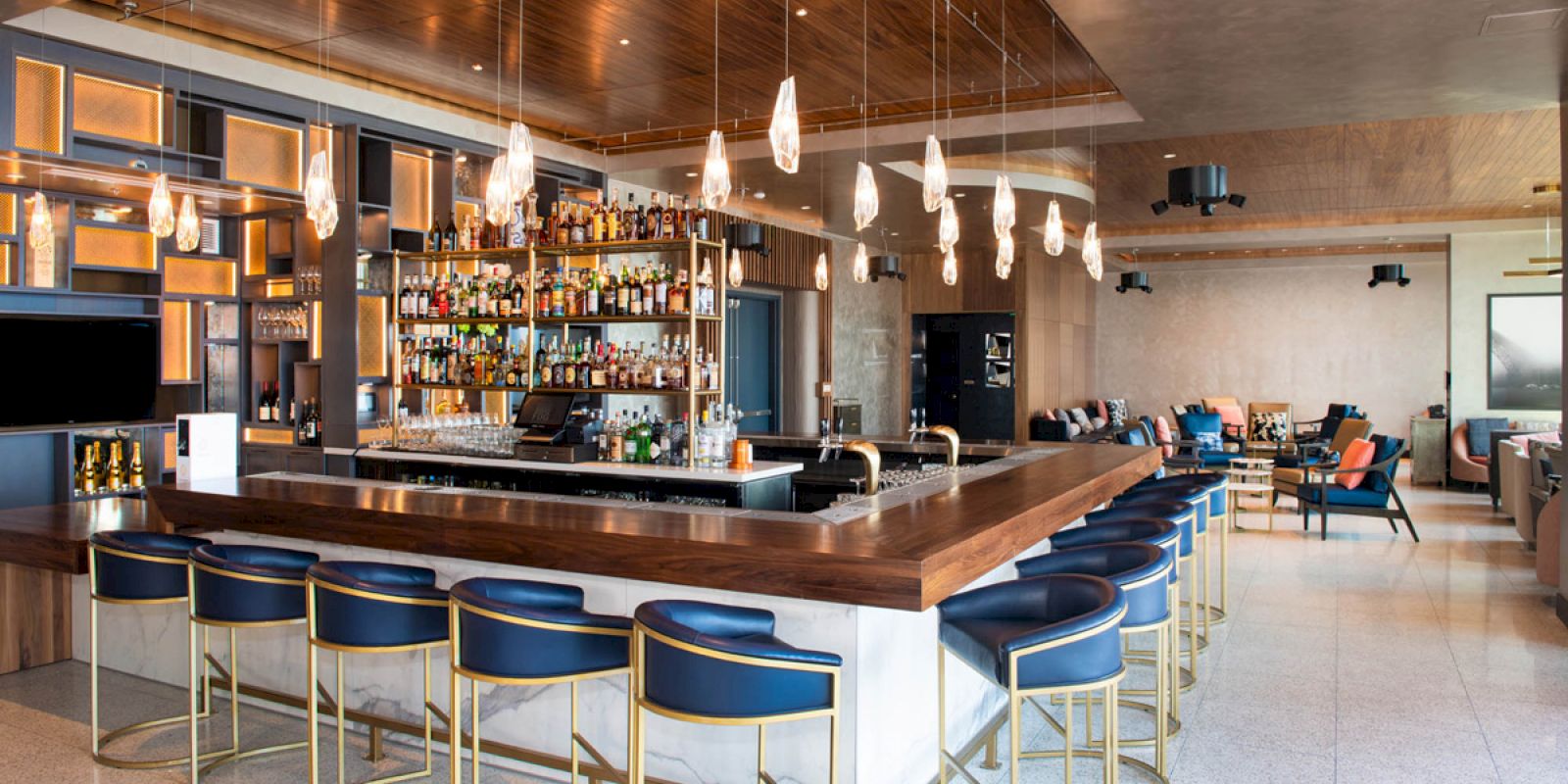 A modern bar with blue stools, pendant lights, and well-stocked shelves, adjacent to a lounge area with seating.