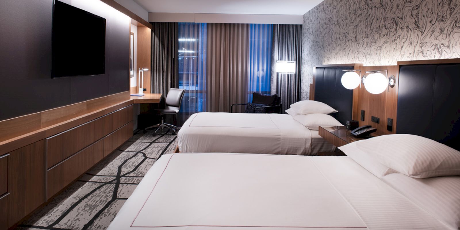 A modern hotel room with two beds, a desk, chair, TV, and a patterned wall with lamps, creating a neat, welcoming atmosphere.