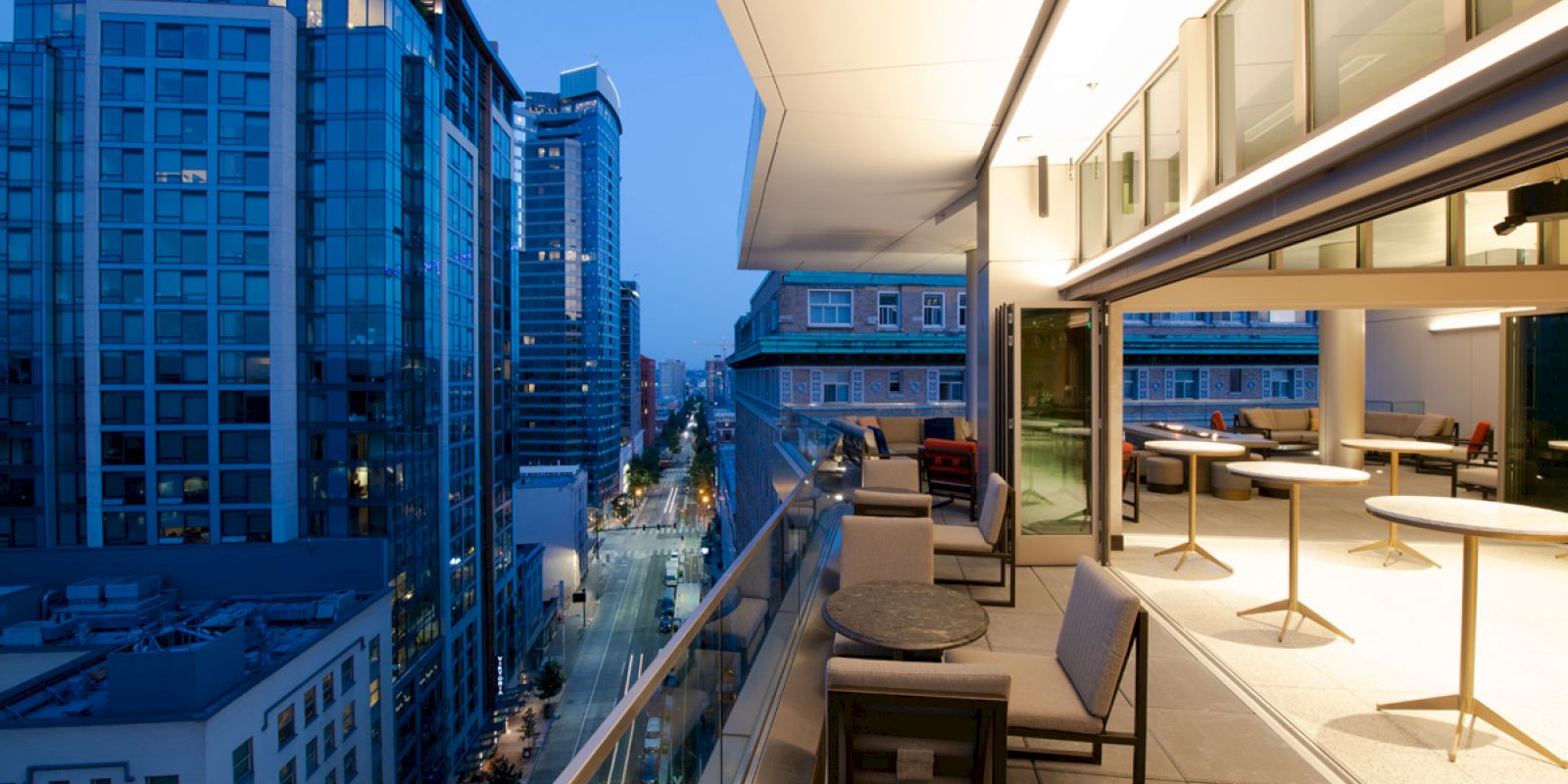 A modern balcony overlooks a cityscape at twilight, with seating and open indoor space visible, surrounded by tall buildings.