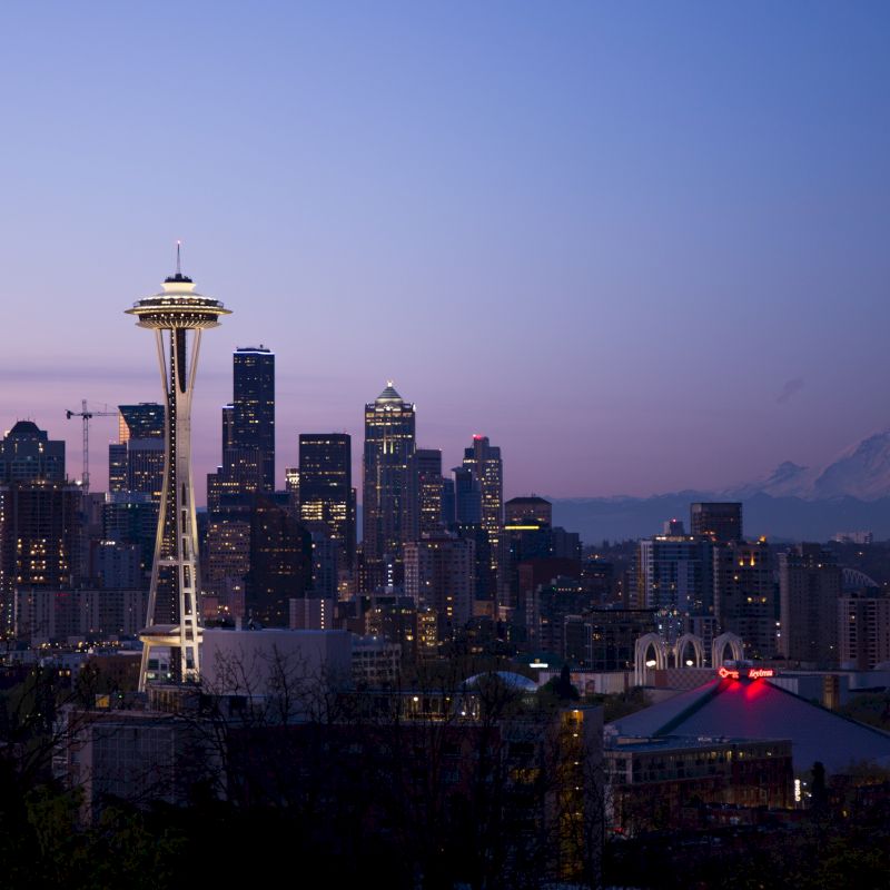 Seattle skyline at dusk with the Space Needle, city buildings, and Mount Rainier in the background, under a clear blue-purple sky.