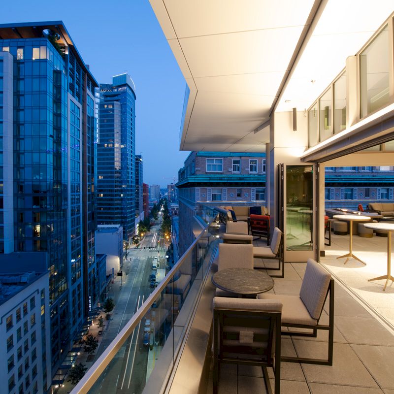 The image shows a modern building with a balcony overlooking a cityscape with tall buildings and a street at twilight.