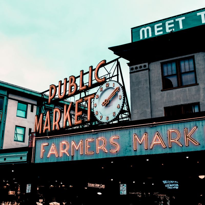 The image shows a public market with neon signs reading "Public Market" and "Farmers Market," featuring an iconic clock and urban buildings.