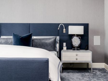 A modern bedroom features a blue and white color scheme, with a neatly made bed, nightstand, lamp, and wall-mounted reading light.