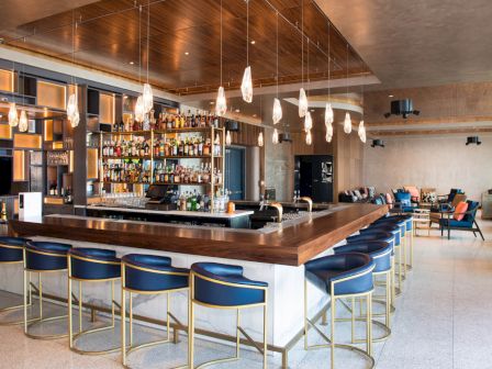 A modern bar with a wooden counter and blue chairs, featuring pendant lights and a fully stocked back bar in a spacious room.