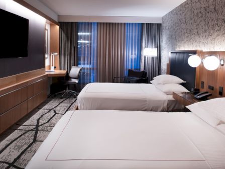 The image shows a modern hotel room with two beds, a TV, a desk with a chair, ambient lighting, and patterned carpet and wallpaper.