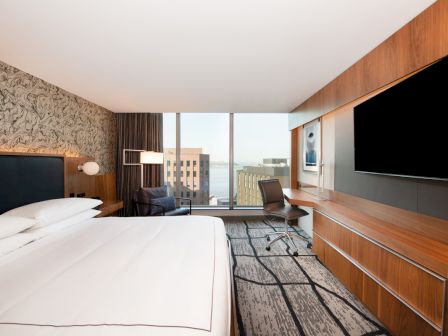 A modern hotel room with a large bed, desk, chair, TV, and window view of city buildings and water in the background.