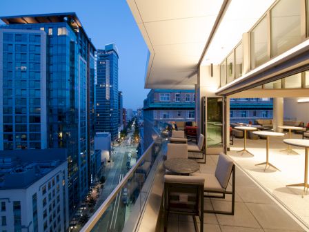 A modern city rooftop terrace with seating overlooks a street lined with tall buildings at dusk.