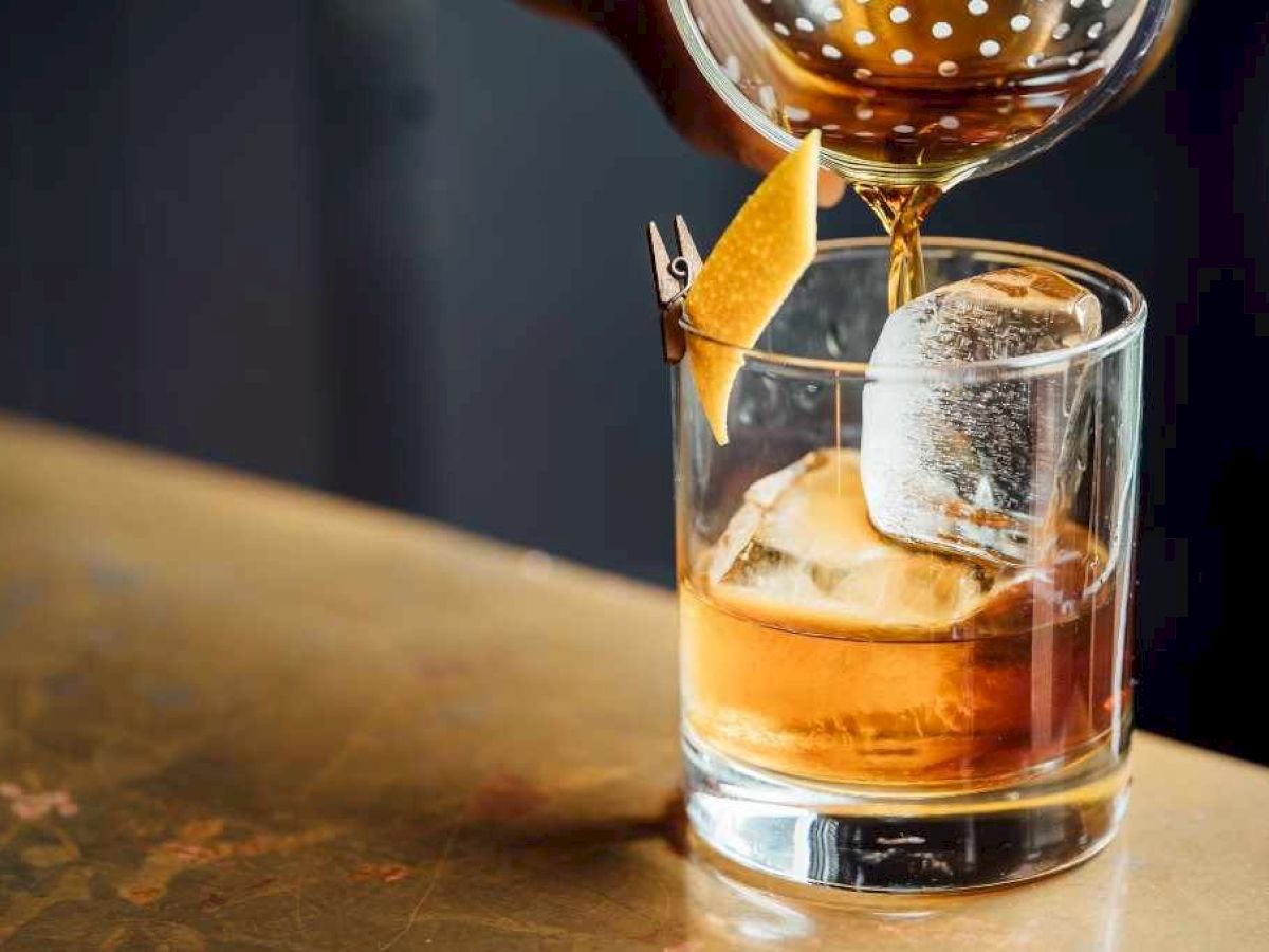 A drink being poured into a glass with large ice cubes and a citrus twist garnish.