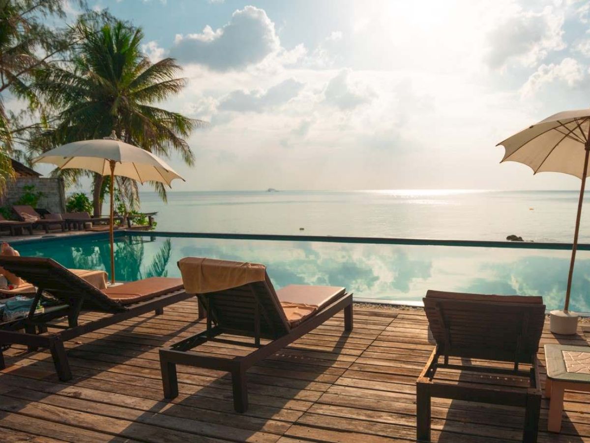 Beachside pool scene with lounge chairs, umbrellas, and palm trees overlooking a calm ocean, under a bright sky.